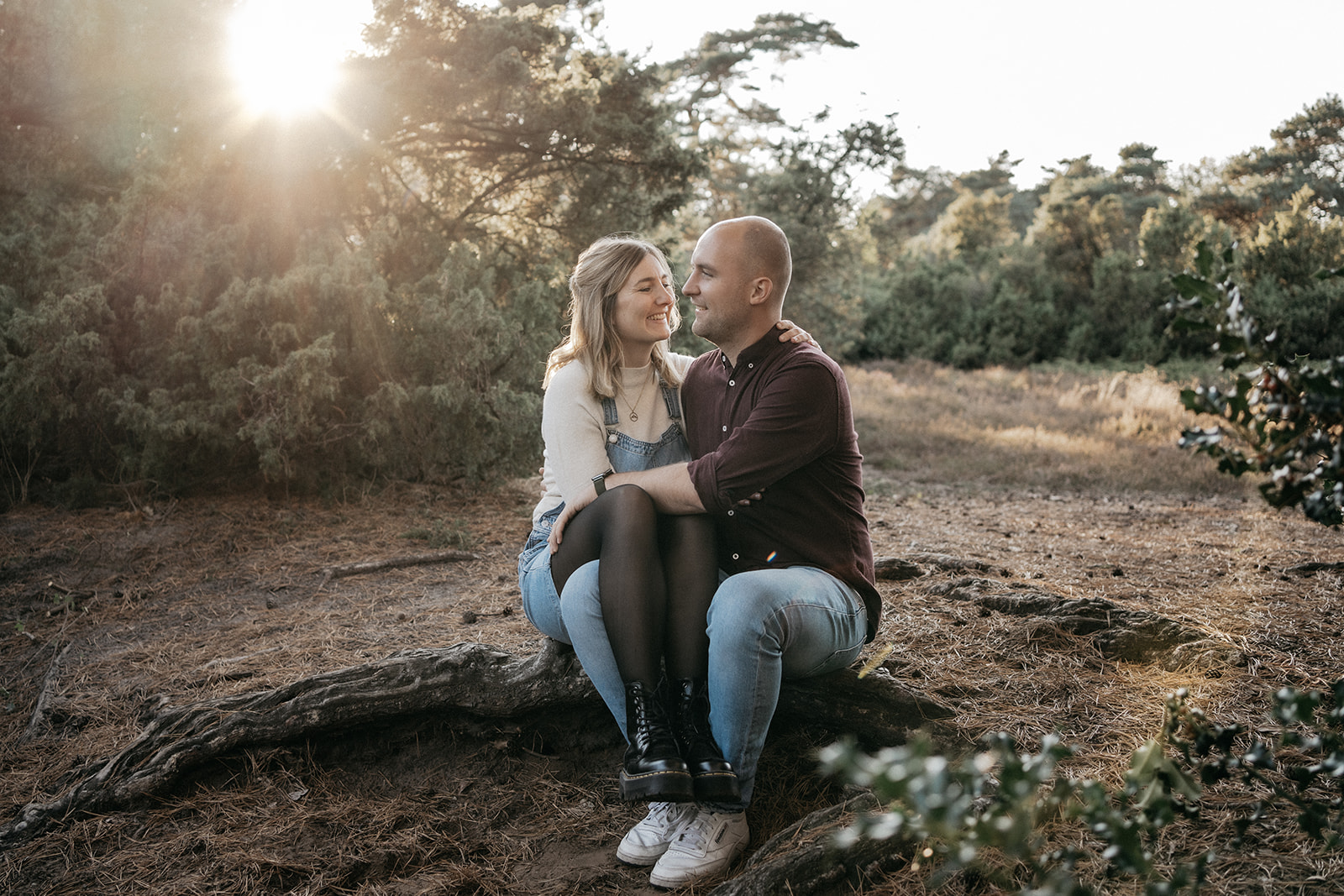 erliebtes Paar beim Paarshooting in Ahaus, sitzt lachend auf einem Baumstamm in der Natur bei Sonnenlicht.