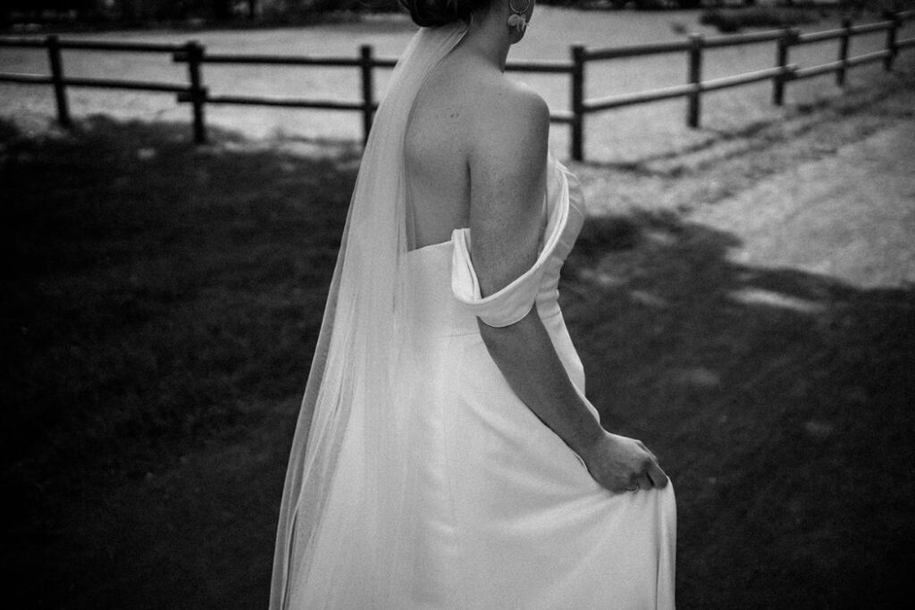 Braut mit Schleier hält ihr Hochzeitskleid im Freien und geht über eine Wiese – Hochzeitsfotograf Ahaus