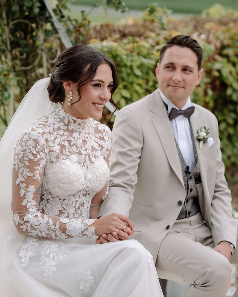 Braut und Bräutigam sitzen bei der Trauung im Grünen und halten sich an den Händen – Hochzeitsfotograf Ahaus