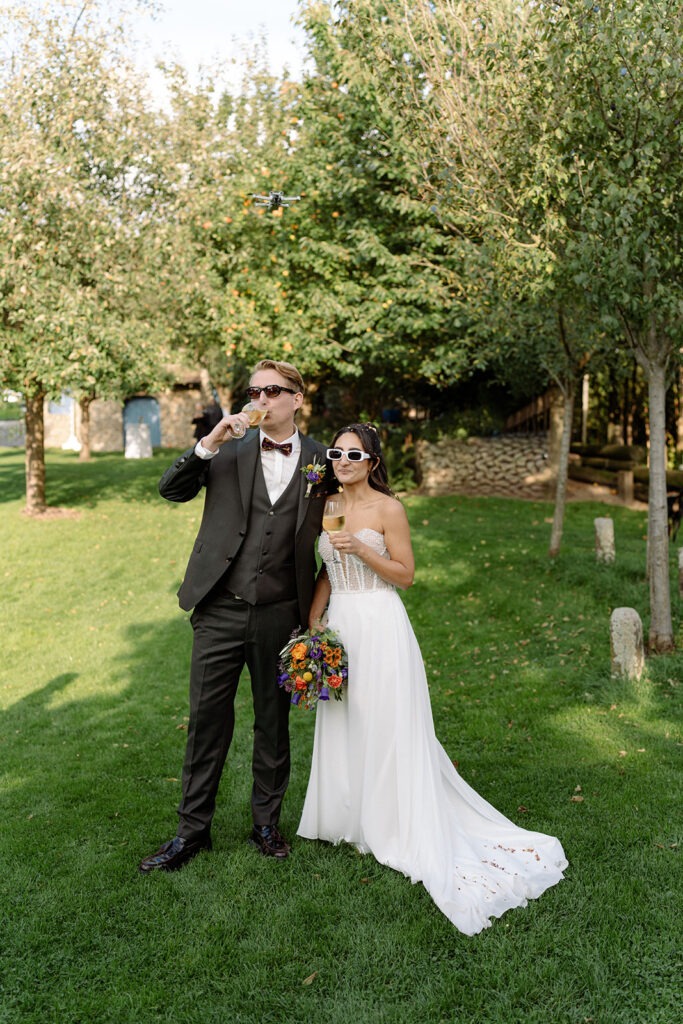 Braut und Bräutigam stoßen im Grünen mit Sekt an und tragen Sonnenbrillen – Hochzeitsfotograf Ahaus
