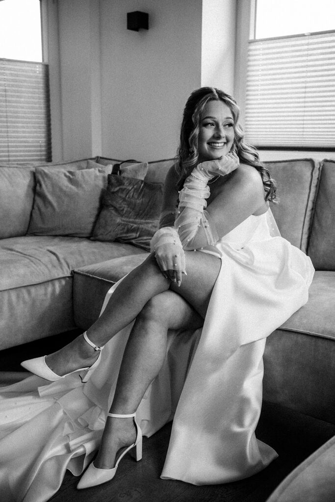Lächelnde Braut im eleganten Hochzeitskleid sitzt beim Getting Ready auf einem Sofa – Hochzeitsfotograf Ahaus